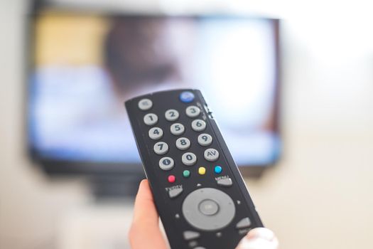 Holding a TV remote control in the hand, foreground, tv in the blurry background. Streaming.
