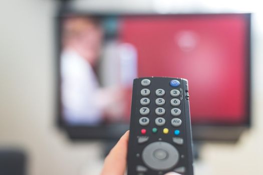 Holding a TV remote control in the hand, foreground, tv in the blurry background. Streaming.