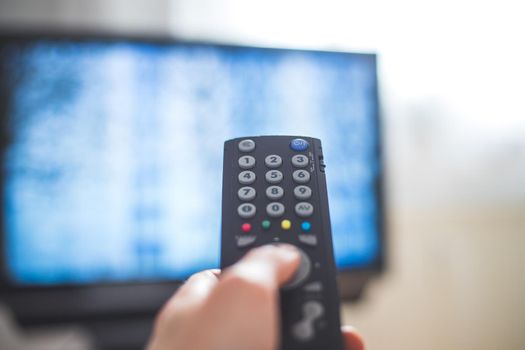 Holding a TV remote control in the hand, foreground, tv in the blurry background. Streaming.
