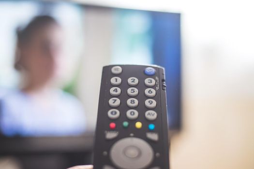 Holding a TV remote control in the hand, foreground, tv in the blurry background. Streaming.