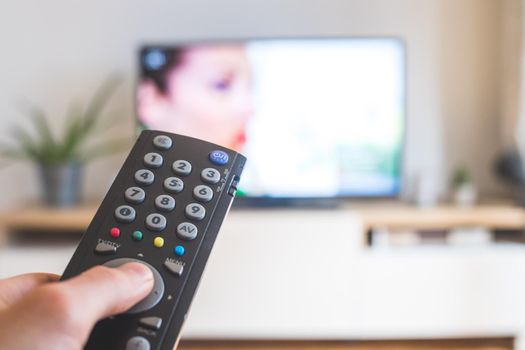 Holding a TV remote control in the hand, foreground, tv in the blurry background. Streaming.