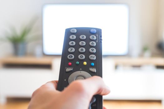 Holding a TV remote control in the hand, foreground, tv in the blurry background. Streaming.