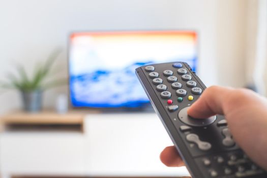 Holding a TV remote control in the hand, foreground, tv in the blurry background. Streaming.