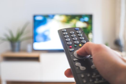 Holding a TV remote control in the hand, foreground, tv in the blurry background. Streaming.