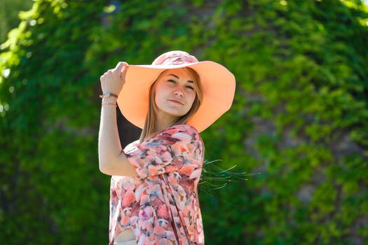 A charming girl in a light summer sundress and a pareo hat is walking in a green park. Enjoys warm sunny summer days.