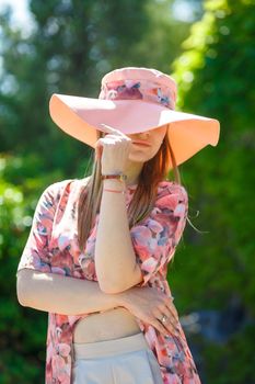 A charming girl in a light summer sundress and a pareo hat is walking in a green park. Enjoys warm sunny summer days.