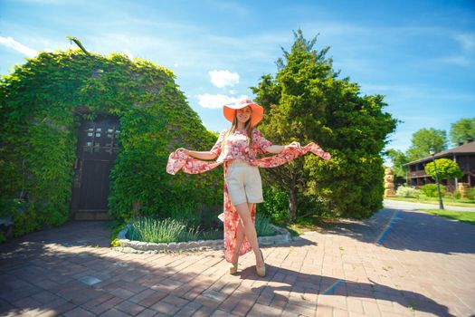 A charming girl in a light summer sundress and a pareo hat is walking in a green park. Enjoys warm sunny summer days.