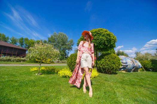 A charming girl in a light summer sundress and a pareo hat is walking in a green park. Enjoys warm sunny summer days.