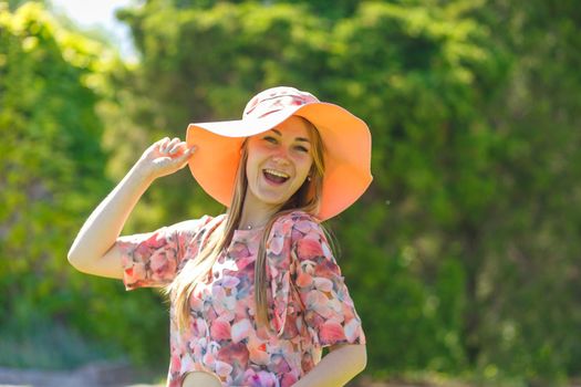 A charming girl in a light summer sundress and a pareo hat is walking in a green park. Enjoys warm sunny summer days.