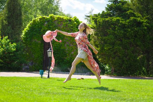 A charming girl in a light summer sundress and a pareo hat is walking in a green park. Enjoys warm sunny summer days.