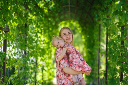 A charming girl in a light summer sundress walks in a green park with her little daughter, holding her in her arms. Enjoys warm sunny summer days.