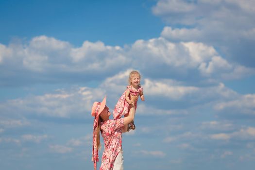 A charming girl in a light summer sundress walks on the sandy beach with her little daughter. Enjoys warm sunny summer days.