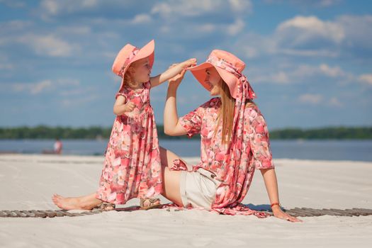 A little girl with her mother in matching beautiful sundresses plays in the sand on the beach. Stylish family look.