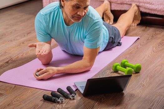 50-year-old man performs exercises while lying on the rug at home, looking at the computer. During a pandemic, a person trains in an apartment via the Internet.