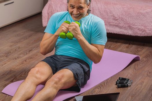 50-year-old man performs exercises while lying on the rug at home, looking at the computer. During a pandemic, a person trains in an apartment via the Internet.