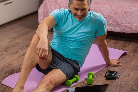 50-year-old man performs exercises while lying on the rug at home, looking at the computer. During a pandemic, a person trains in an apartment via the Internet.