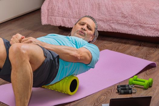 50-year-old man performs exercises while lying on the rug at home, looking at the computer. During a pandemic, a person trains in an apartment via the Internet.