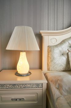 table lamp on a bedside table in a bedroom with gray walls near the bed is lit with yellow light