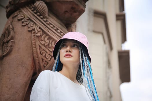 Happy stylish young hipster woman with long blue hair pink jacket, hat on the street in urban city.