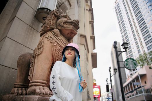 Happy stylish young hipster woman with long blue hair pink jacket, hat on the street in urban city.