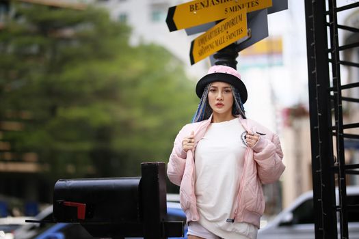 Happy stylish young hipster woman with long blue hair pink jacket, hat on the street in urban city.