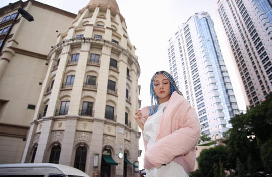 Happy stylish young hipster woman with long blue hair pink jacket, hat on the street in urban city.