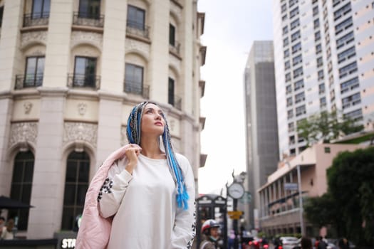 Happy stylish young hipster woman with long blue hair pink jacket, hat on the street in urban city.