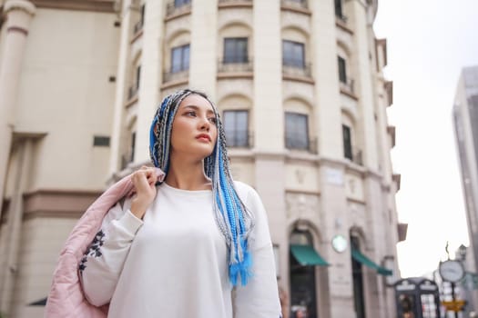 Happy stylish young hipster woman with long blue hair pink jacket, hat on the street in urban city.