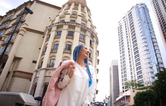 Happy stylish young hipster woman with long blue hair pink jacket, hat on the street in urban city.