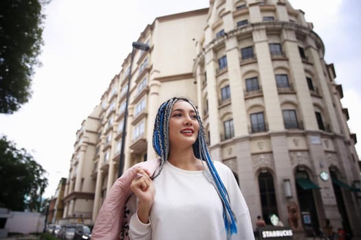 Happy stylish young hipster woman with long blue hair pink jacket, hat on the street in urban city.