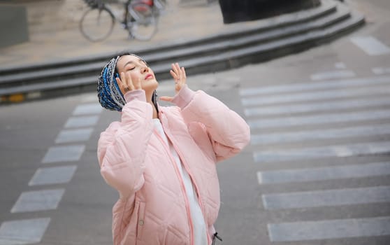 Happy stylish young hipster woman with long blue hair pink jacket, hat on the street in urban city.