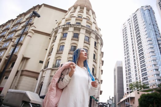 Happy stylish young hipster woman with long blue hair pink jacket, hat on the street in urban city.