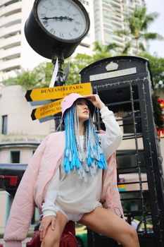 Happy stylish young hipster woman with long blue hair pink jacket, hat on the street in urban city.