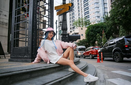 Happy stylish young hipster woman with long blue hair pink jacket, hat on the street in urban city.