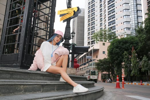 Happy stylish young hipster woman with long blue hair pink jacket, hat on the street in urban city.