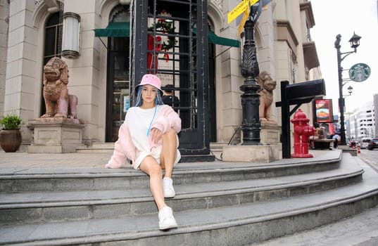 Happy stylish young hipster woman with long blue hair pink jacket, hat on the street in urban city.