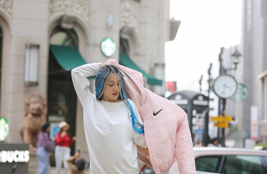 Happy stylish young hipster woman with long blue hair pink jacket, hat on the street in urban city.