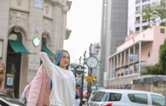 Happy stylish young hipster woman with long blue hair pink jacket, hat on the street in urban city.