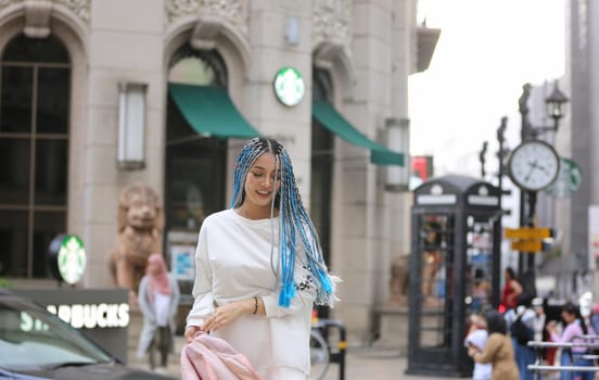 Happy stylish young hipster woman with long blue hair pink jacket, hat on the street in urban city.