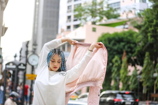 Happy stylish young hipster woman with long blue hair pink jacket, hat on the street in urban city.