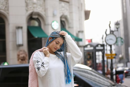 Happy stylish young hipster woman with long blue hair pink jacket, hat on the street in urban city.