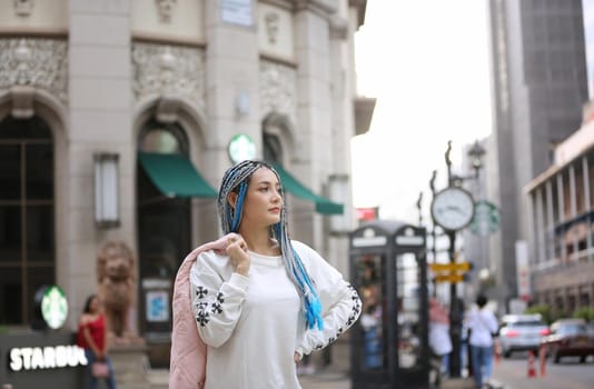 Happy stylish young hipster woman with long blue hair pink jacket, hat on the street in urban city.