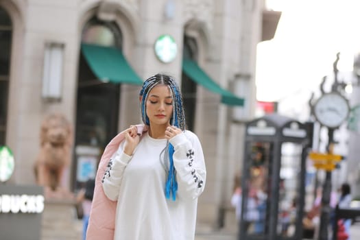 Happy stylish young hipster woman with long blue hair pink jacket, hat on the street in urban city.