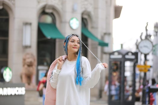 Happy stylish young hipster woman with long blue hair pink jacket, hat on the street in urban city.