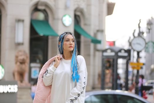 Happy stylish young hipster woman with long blue hair pink jacket, hat on the street in urban city.