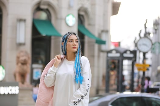 Happy stylish young hipster woman with long blue hair pink jacket, hat on the street in urban city.