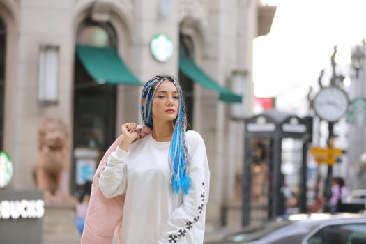 Happy stylish young hipster woman with long blue hair pink jacket, hat on the street in urban city.