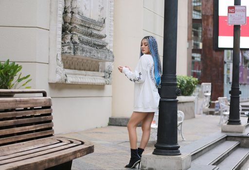 Happy stylish young hipster woman with long blue hair pink jacket, hat on the street in urban city.