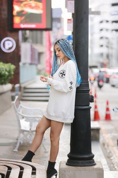 Happy stylish young hipster woman with long blue hair pink jacket, hat on the street in urban city.