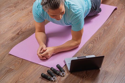 50-year-old man performs exercises while lying on the rug at home, looking at the computer. During a pandemic, a person trains in an apartment via the Internet.